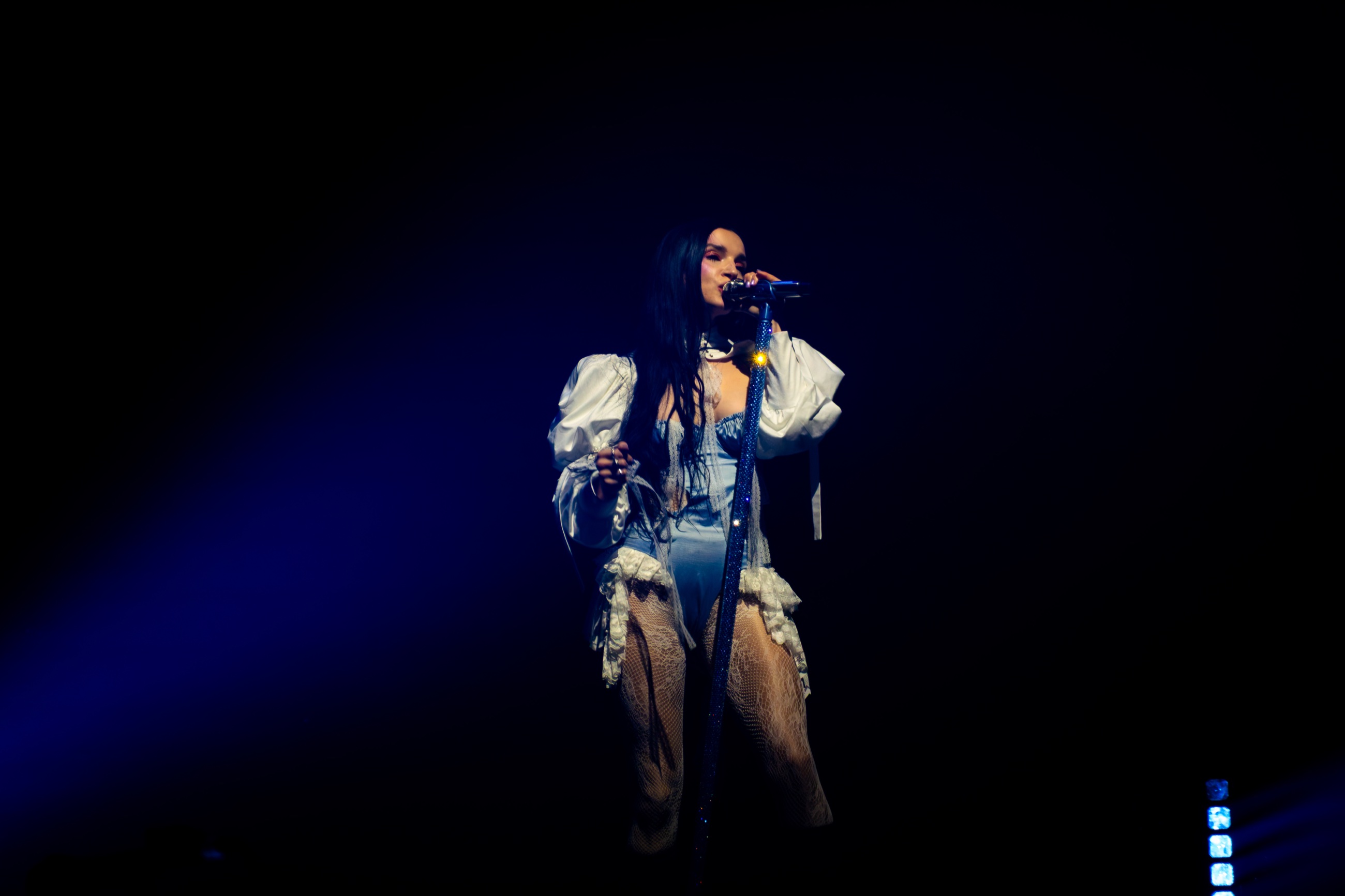 Poppy singing at a crystal-covered microphone stand beneath deep blue stage light