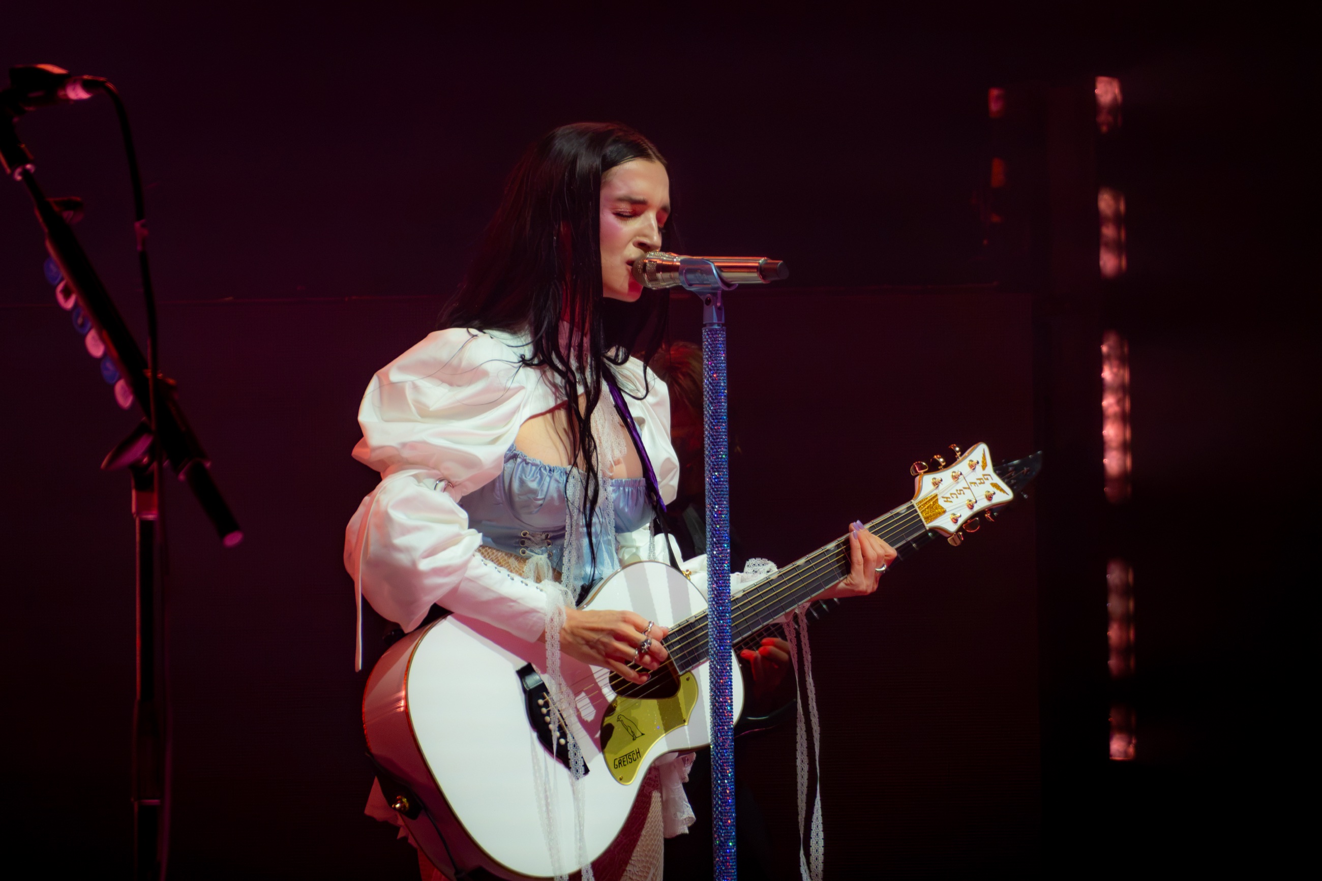 Poppy plays a white guitar and sings beneath warm red stage light